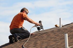Homeowner examining dark roof stains and green moss near shingles with a flashlight and ladder.
