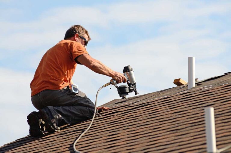 Homeowner examining dark roof stains and green moss near shingles with a flashlight and ladder.